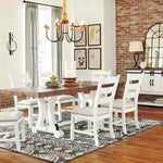 A dining room scene showing a white wooden dining table with six matching ladder-back chairs arranged on a patterned rug. The room has exposed brick walls, hardwood floors, a chandelier, framed wall art, and a white sideboard cabinet with lamps.
