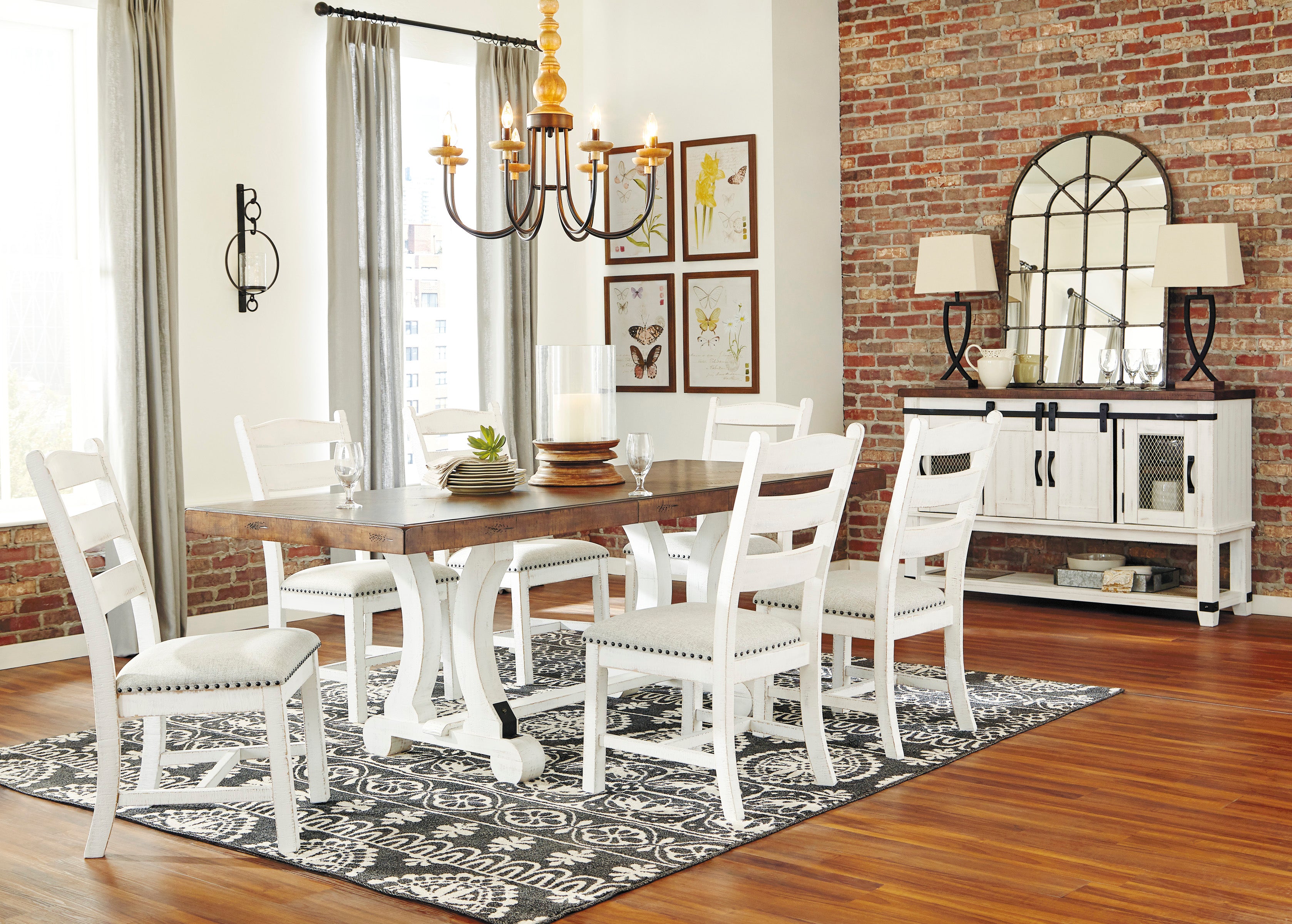 A dining room scene showing a white wooden dining table with six matching ladder-back chairs arranged on a patterned rug. The room has exposed brick walls, hardwood floors, a chandelier, framed wall art, and a white sideboard cabinet with lamps.