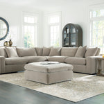 A light beige sectional sofa with matching throw pillows and a matching ottoman, arranged in a bright, modern living room with wood floors, side tables, lamps, and a large patterned area rug.