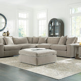 A light beige sectional sofa with matching throw pillows and a matching ottoman, arranged in a bright, modern living room with wood floors, side tables, lamps, and a large patterned area rug.