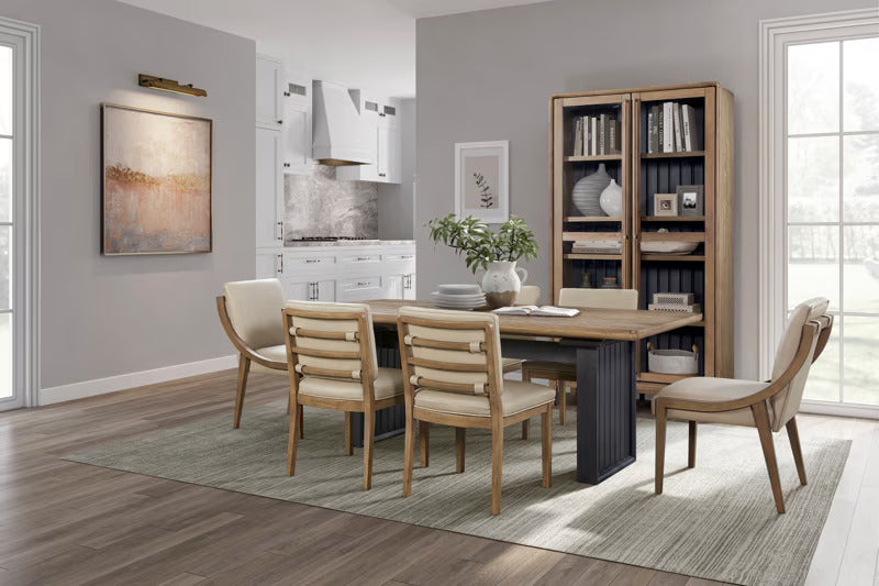 Modern dining room with wooden table and chairs, bookshelf, and kitchen in the background.
