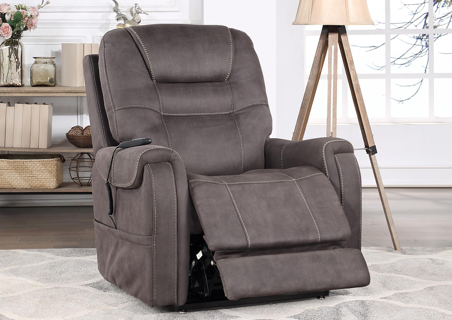 A gray-brown power recliner chair with a footrest extended, shown in a styled living room setting with a tripod floor lamp, shelving, and a window.