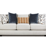 A three-seat upholstered sofa in a light warm gray or off-white fabric, styled with five decorative throw pillows including two navy blue velvet pillows, two patterned white pillows, and one tan striped pillow, shown against a plain white background.