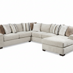 A light beige fabric sectional sofa with a chaise lounge on the right side, styled with multiple throw pillows in neutral tones, set against a plain white background.
