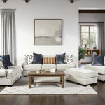 A staged living room featuring a light beige sectional sofa with matching ottoman, a wooden coffee table, patterned throw pillows, and a large framed abstract print on a white wall.