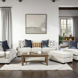 A staged living room featuring a light beige sectional sofa with matching ottoman, a wooden coffee table, patterned throw pillows, and a large framed abstract print on a white wall.
