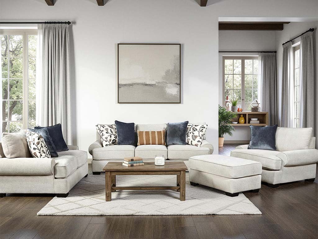 A staged living room featuring a light beige sectional sofa with matching ottoman, a wooden coffee table, patterned throw pillows, and a large framed abstract print on a white wall.
