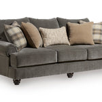 A three-seater sofa upholstered in textured gray fabric, styled with multiple throw pillows in shades of gray, beige, and brown, set against a plain white background.