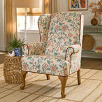 A classic wingback armchair with a floral upholstery pattern in orange, green, and cream, featuring brown wooden legs, set in a beautifully decorated living room next to a woven side table.