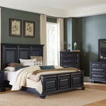 A 7-piece bedroom furniture set in a dark charcoal gray finish, including a bed frame with a paneled headboard, a nightstand, a dresser, a chest, and a matching mirror, styled in a modern bedroom with dark gray walls and beige curtains.