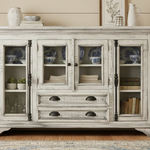 A distressed white wooden sideboard cabinet with glass doors and drawers, styled in a rustic room setting with decorative ceramic vases, books, and plants.