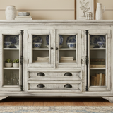 A distressed white wooden sideboard cabinet with glass doors and drawers, styled in a rustic room setting with decorative ceramic vases, books, and plants.