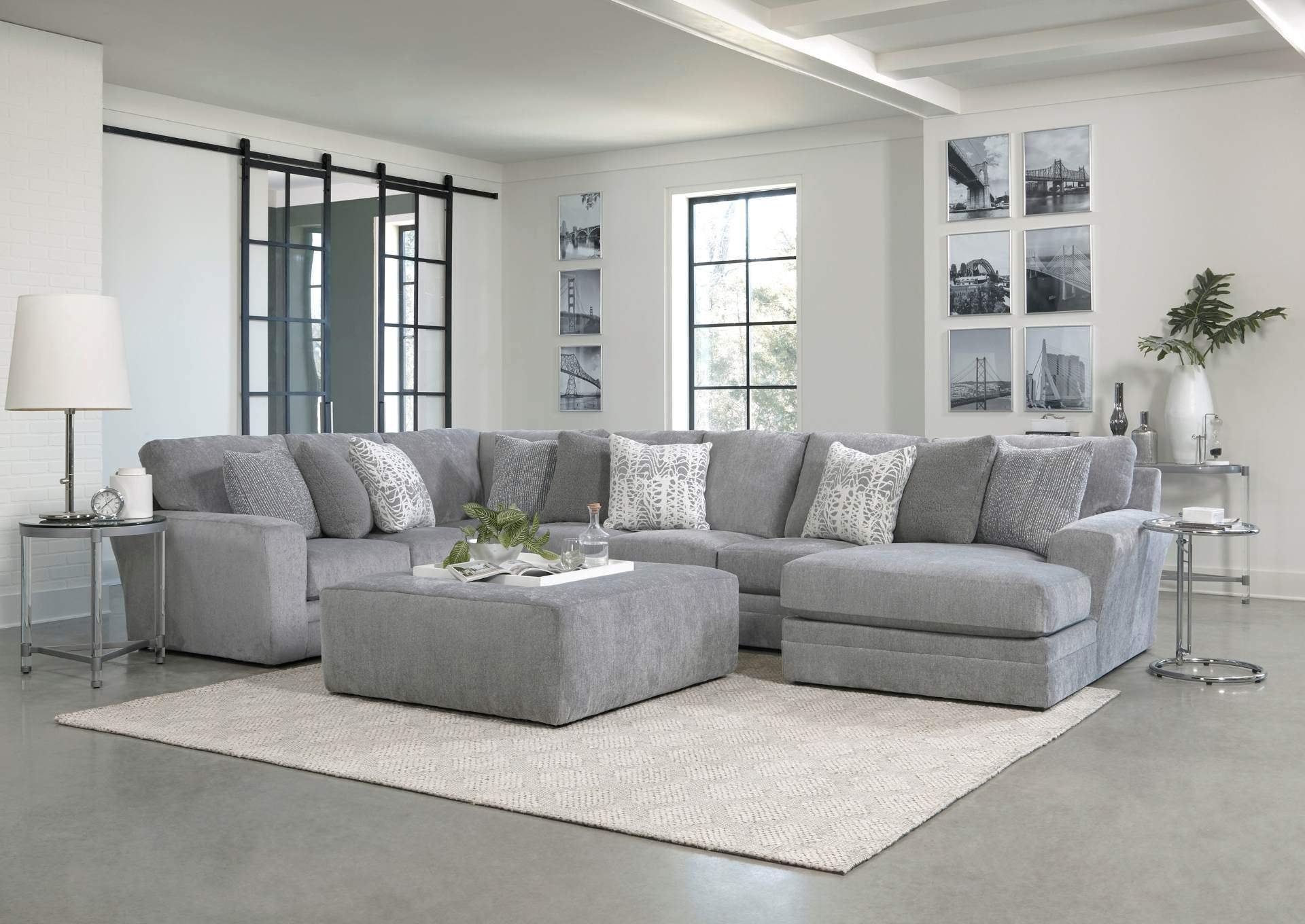 A light gray sectional sofa with matching ottoman and decorative pillows in a bright, modern living room with white walls and large windows.