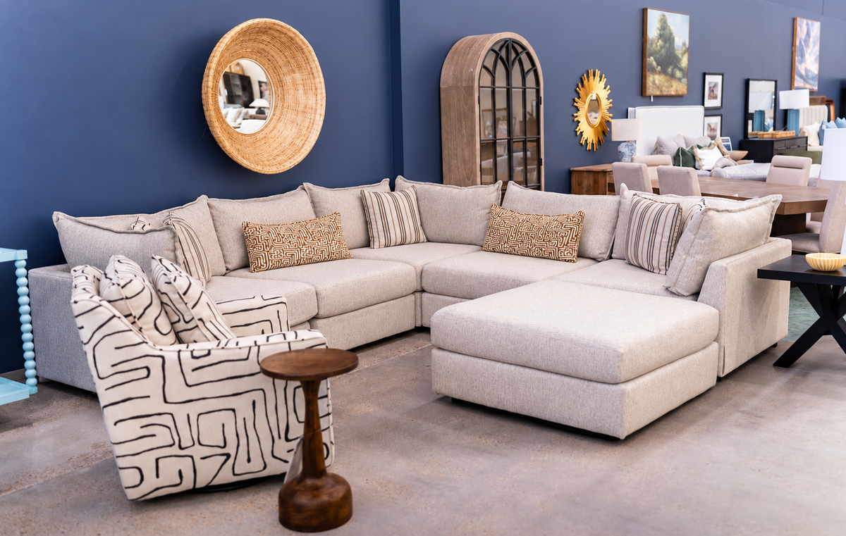 A light beige modular sectional sofa with an ottoman used as a chaise lounge and striped and textured accent pillows, displayed in a furniture showroom with a blue wall and other decor items.